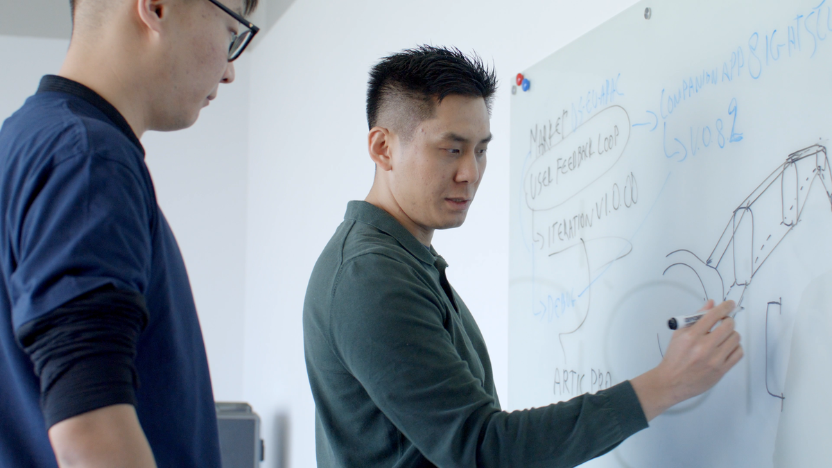 Two engineers reviewing a camera-glasses side profile and technical notes drawn on a whiteboard.