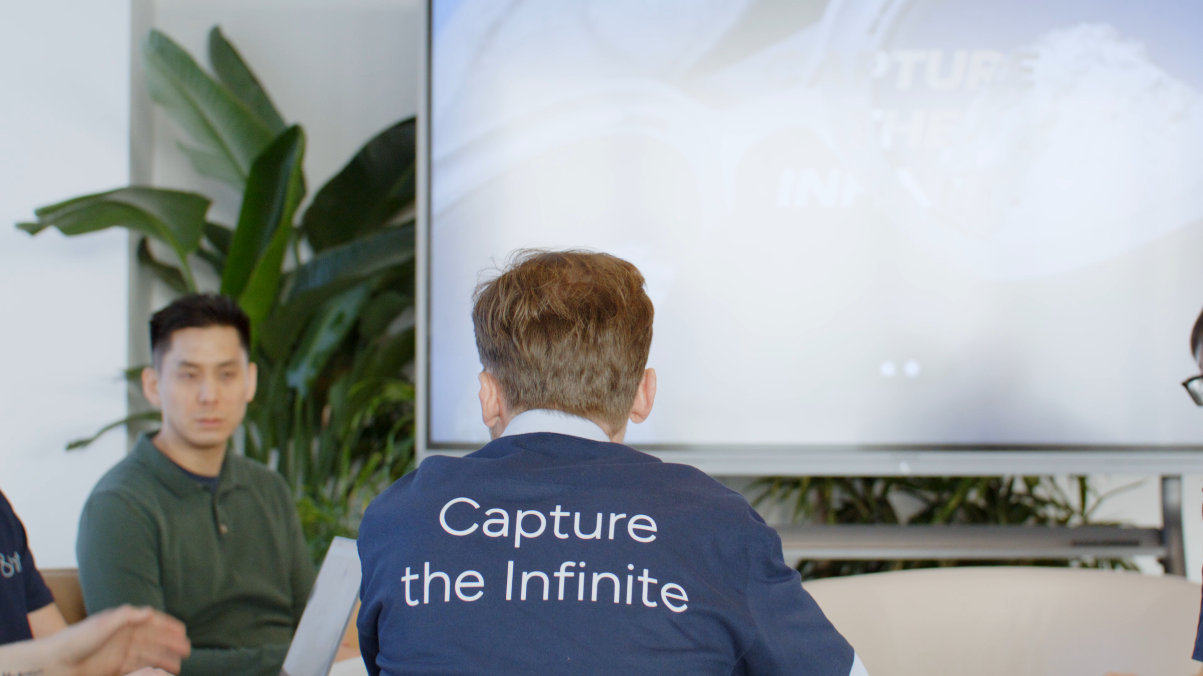 8ight team session viewed behind a shirt reading “Capture the Infinite,” with colleagues and a presentation screen ahead.
