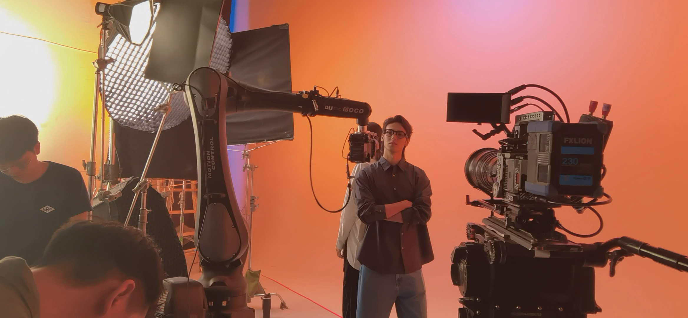 Male model wearing black camera glasses beside cinema cameras on the orange studio set.