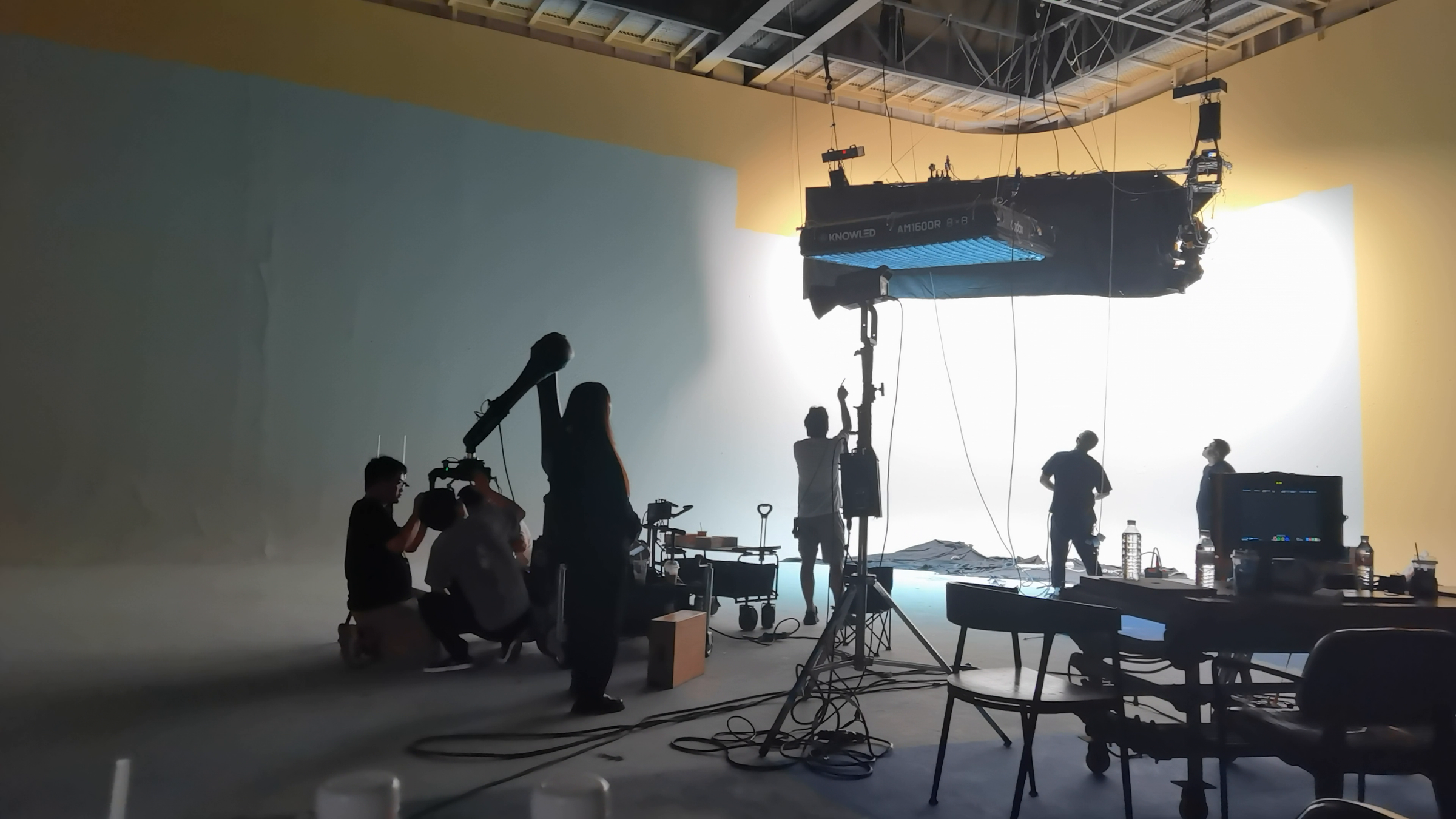 Production crew and model silhouetted beneath a suspended softbox on a large curved studio backdrop.
